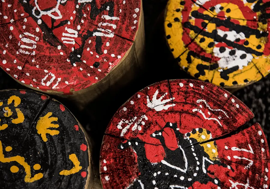 Painted wooden discs featuring Aboriginal‑inspired symbols and patterns in red, yellow, black and white, arranged closely together as examples of children’s creative artwork.