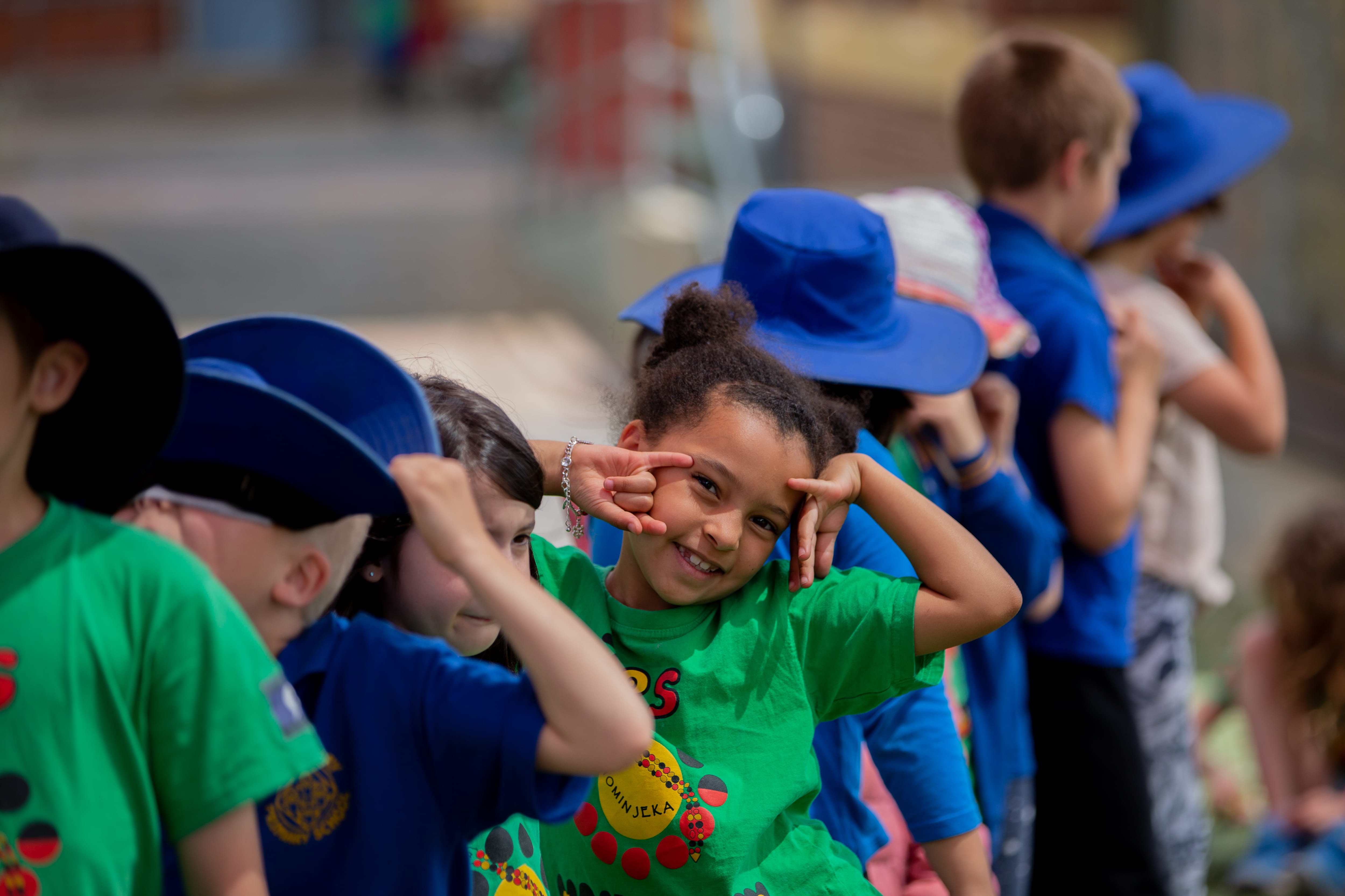 A group of primary school aged students standing in a line. One students is looking at the camera smiling, with her hands raised to her face.